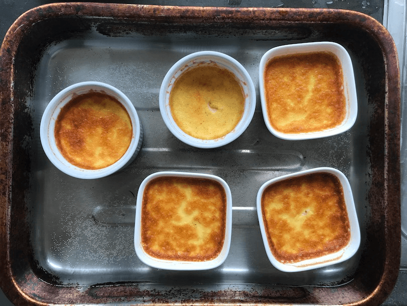 Five ramekins filled with custard, caramelising on top, sitting in an old oven dish.