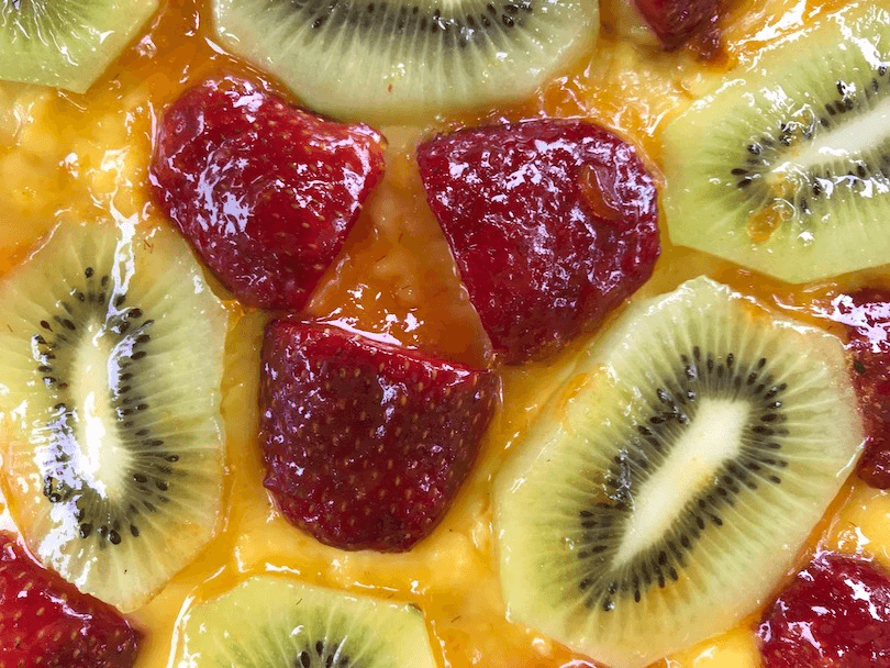 Aerial view of a section of a fruit flan. You see custard underneath, then a layer of sliced strawberries and kiwifruit, all covered in a slick apricot jam glaze.