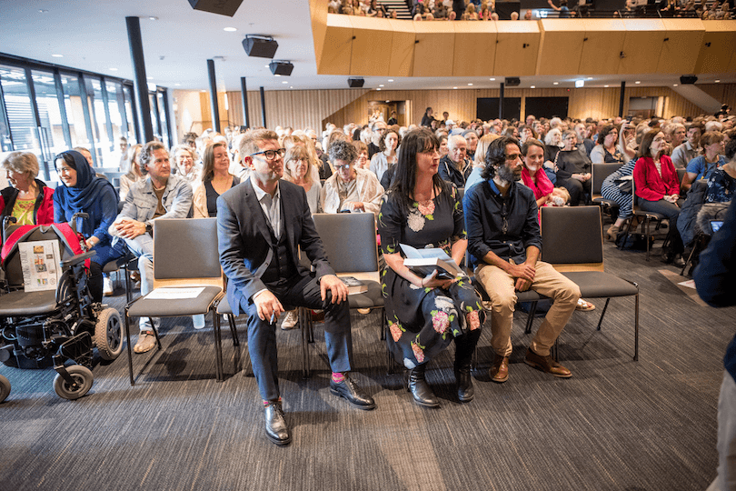A big, high-ceilinged room, packed with people seated in chairs. Three people in front row are all looking eagerly at something off-camera. 