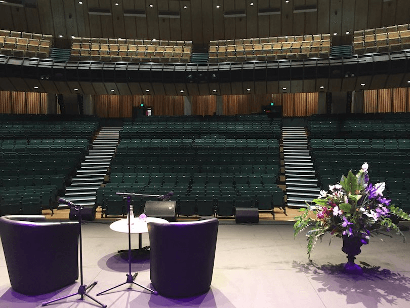 An empty auditorium photographed from the stage, where we see two empty chairs, a small table, and huge bunch of flowers.