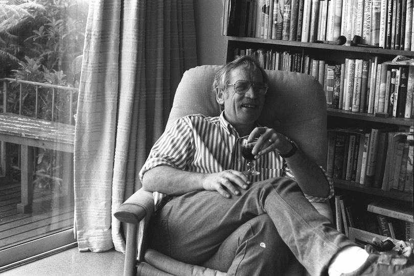 Black and white photo of a NZ lounge or library room, a middle-aged man sitting very comfortably in a chair, red wine in hand, laughing.