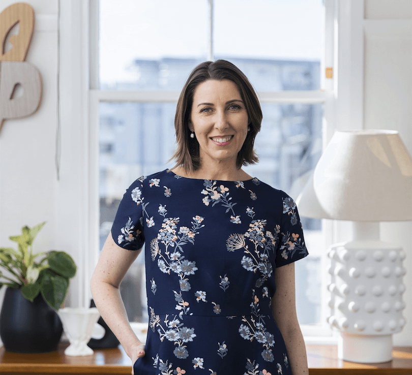 Middle-aged white woman photographed in an airy apartment, houseplant, cool lamp etc. 
