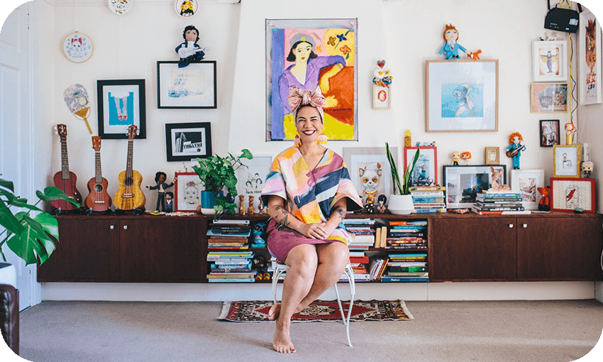 Vibrant photo of a woman sitting at home, in front of a gorgeous bookcase full of musical instruments and books, a wall covered with art. The woman is beaming.