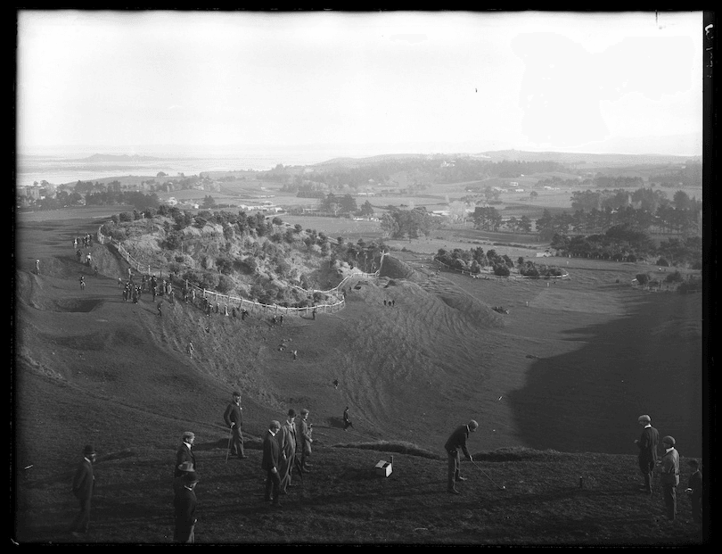 Black and white photo taken from elevated point, looking down at a crater / mountain slopes. A bunch of men are playing golf, bystanders in attendance. 