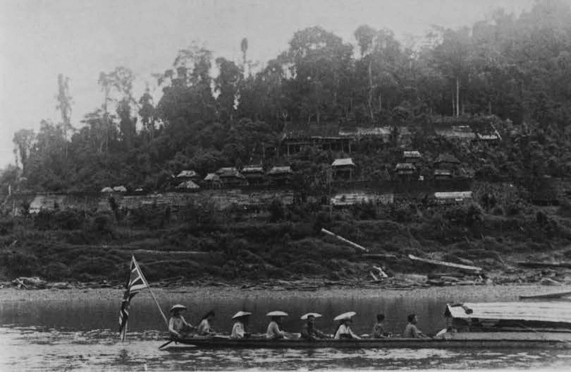 Black and white photo of a tropical river, we're looking across the width of it, foreground dominated by a large canoe with eight people in it. Flag flying at stern. 