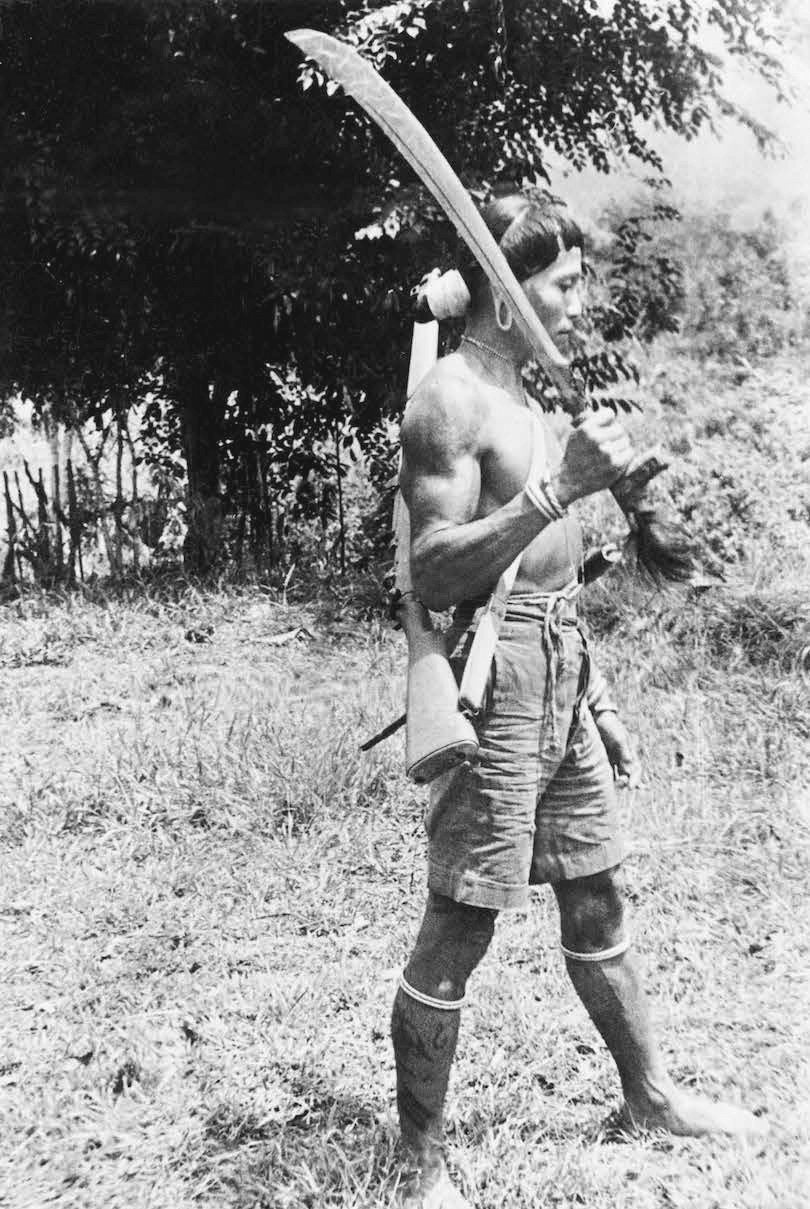 Black and white photograph showing an indigenous man holding a huge blade casually across his shoulder. Photo taken outdoors. 