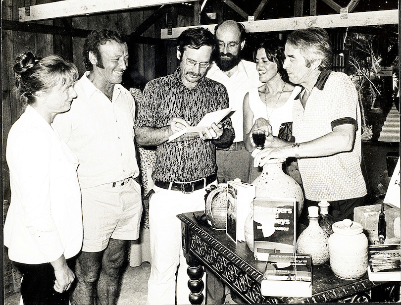 Black and white photo of a group of six adults, focused on man in centre of group who is signing a book. Pottery on table in foreground.