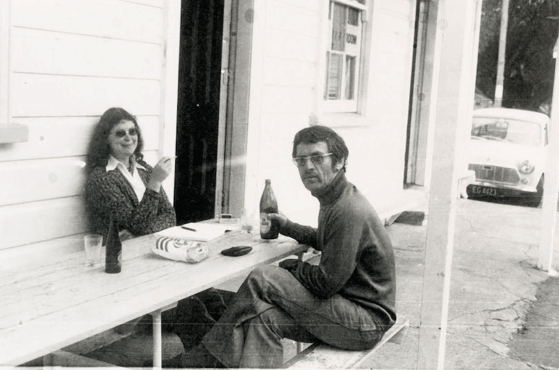 Black and white photo of exterior of a tearooms, a man and woman sitting at wooden table. He's drinking a crate bottle and she's smoking, smiling.
