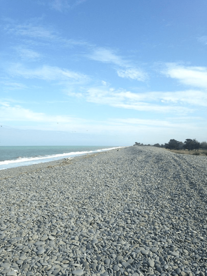 Portrait-shape photograph showing a blue sky with white scuddy clouds, and a stony beach stretching on forever.
