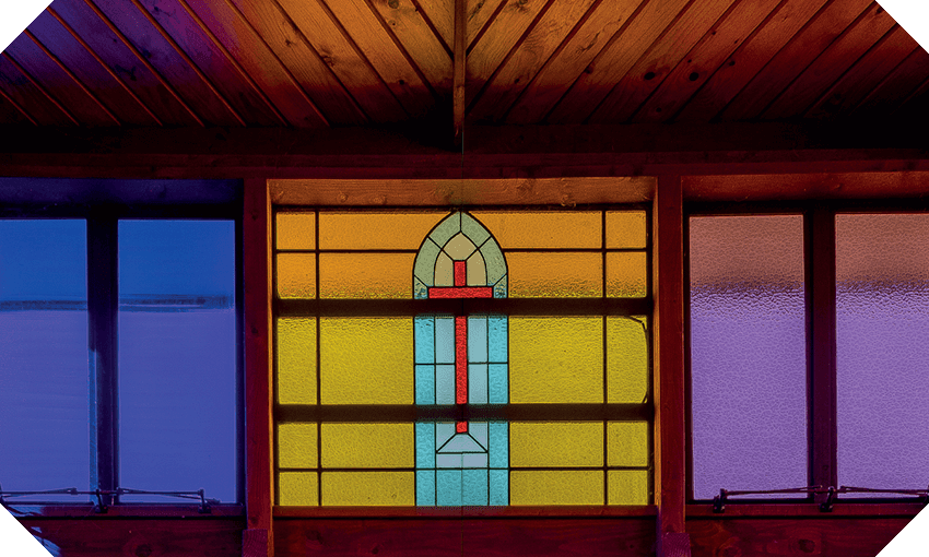 Gorgeous photograph of a stained-glass window, all yellow, purple and blue, with a crucifix picked out in the centre. Wooden ceilings soaring above.