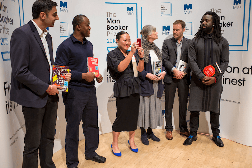 Six writers arranged in front of Man Booker Prize posters.  Photo taken at a candid moment, the smaller woman in the middle (Hanya Yanagihara) takes a selfie. 