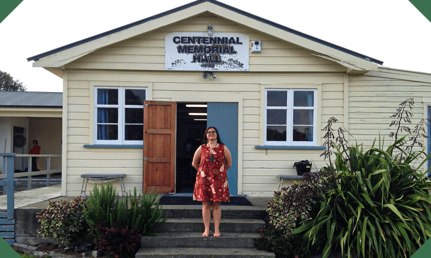 A young Māori woman stands proudly on the steps of a classic Kiwi community hall, harakeke to one side.