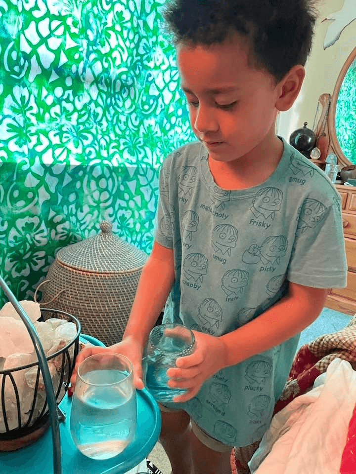 Photo of young boy carefully replacing one glass of water with another, on what looks like a side table. Backdrop of bright turquoise fabric. 