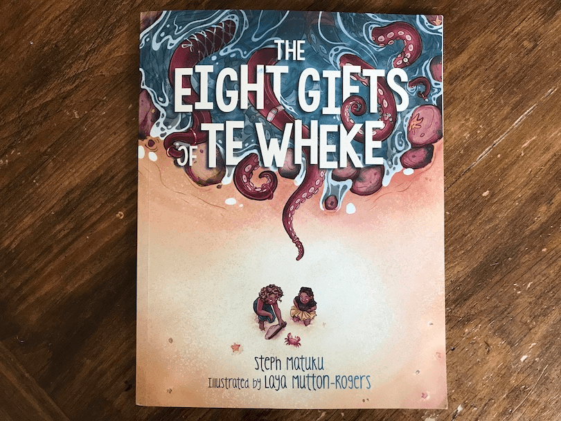 Photograph of picture book showing birds eye view of two children on a beach facing a giant octopus in the water.