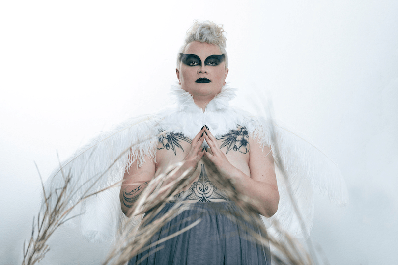 Photograph of a woman dressed magnificently in white-feathered wings and dramatic makeup. 