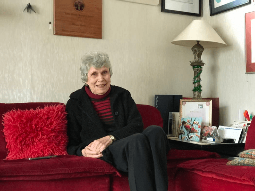 Dame Margaret Sparrow, smiling and sitting on a red couch