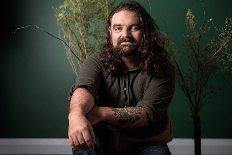 Portrait of a long-haired young Māori man, backdrop is a deep green wall, branches of mānuka arranged behind him.