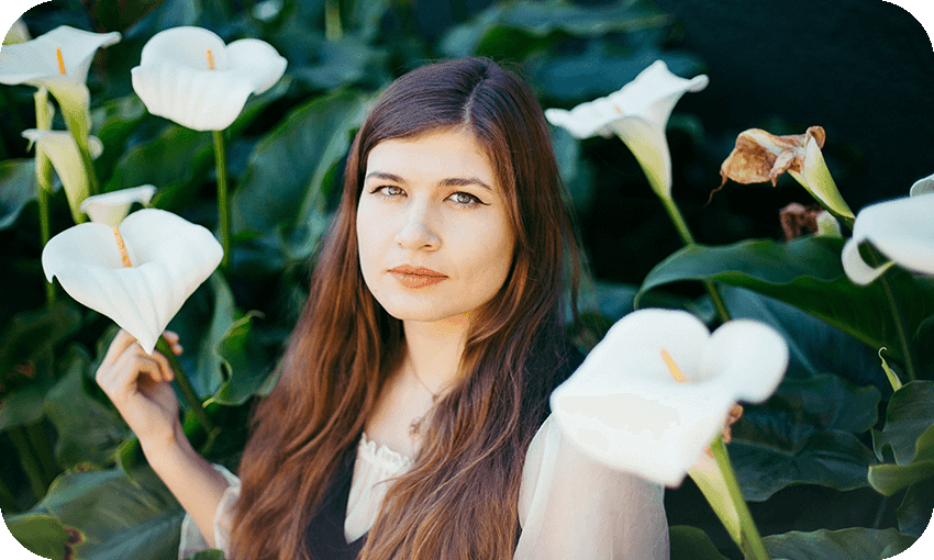 Young woman with long brown hair staring serenely at camera, backdrop of arum lilies.