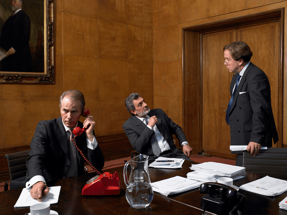 Three older white men in business suits gathered around a boardroom table