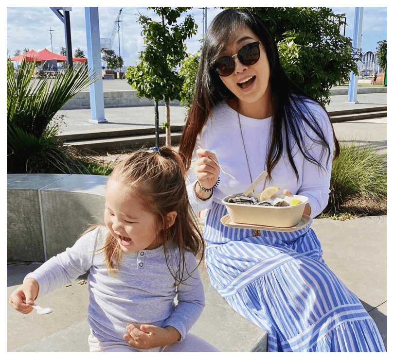 Photo of a mother and daughter perched on a wall, beautiful sunny day, tucking into kai. 