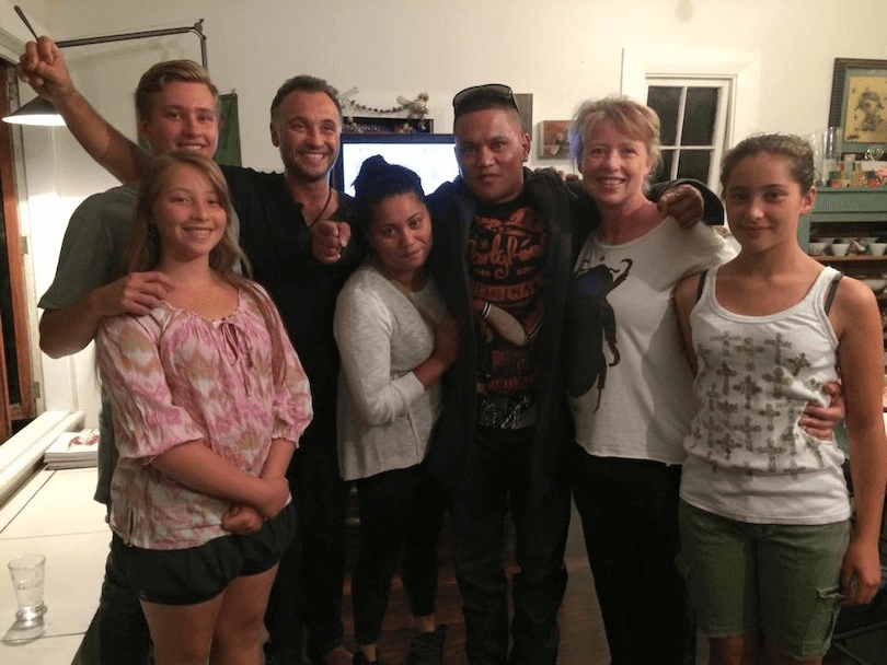 Photo of seven people taken in a living room.  They stand close to each other, arms around each other, there is a sense of excitement and relief. 