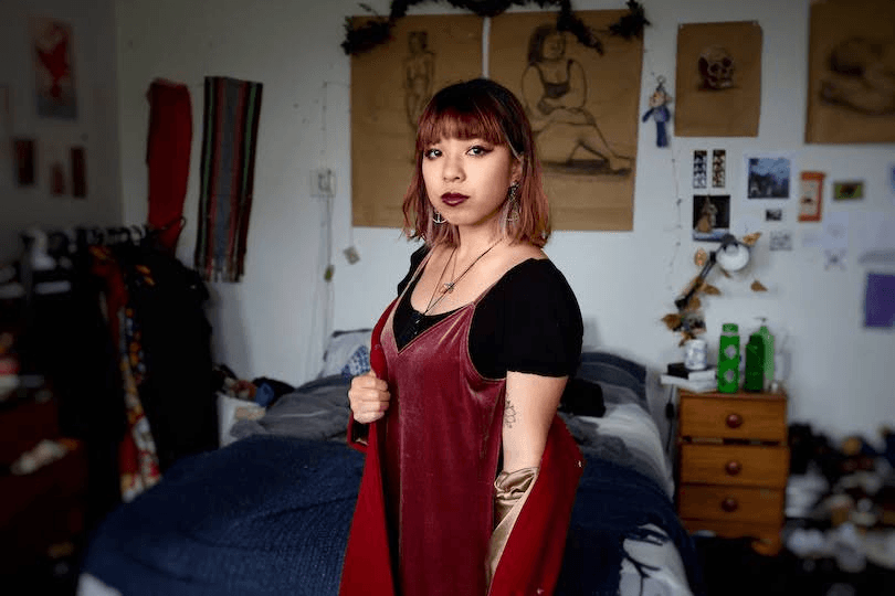 Photo of young woman in her bedroom, standing front and centre, looking straight down the barrel. She wears bronze eye makeup and a velvet dress.
