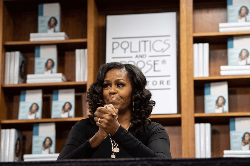 Michelle Obama seated in front of a huge shelf stacked with copies of her memoir. She's clasping her hands together and looking ... pensive, maybe?