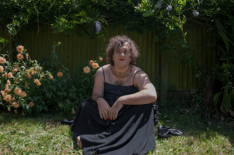 The poet in a garden of orange dahlias and purple wisteria. They're seated and wearing a black maxi dress. 