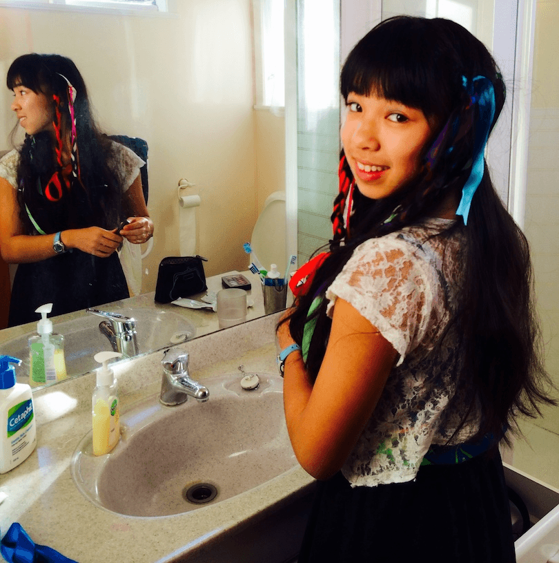 Photo of a teenaged girl standing in front of a bathroom vanity, looking back over her shoulder at the camera. 
