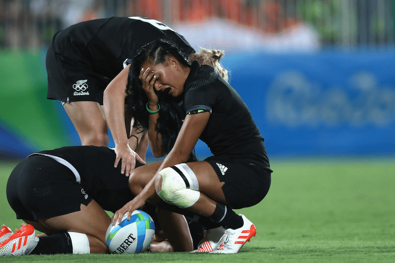 Photograph of three women's rugby players. One is curled up in a ball on the ground, the others are bending to comfort her. They look strong but devastated.