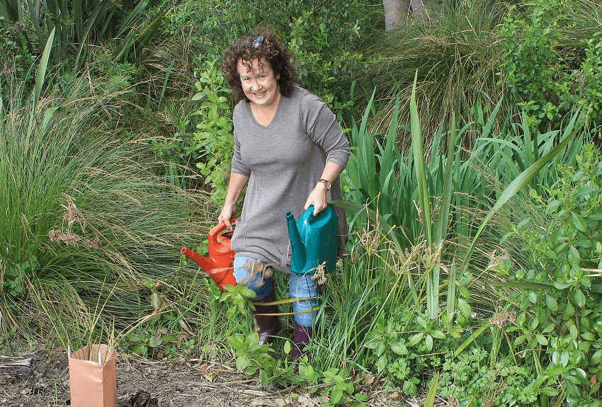 middle aged white woman in grey dress with two watering cans surrounded by trees