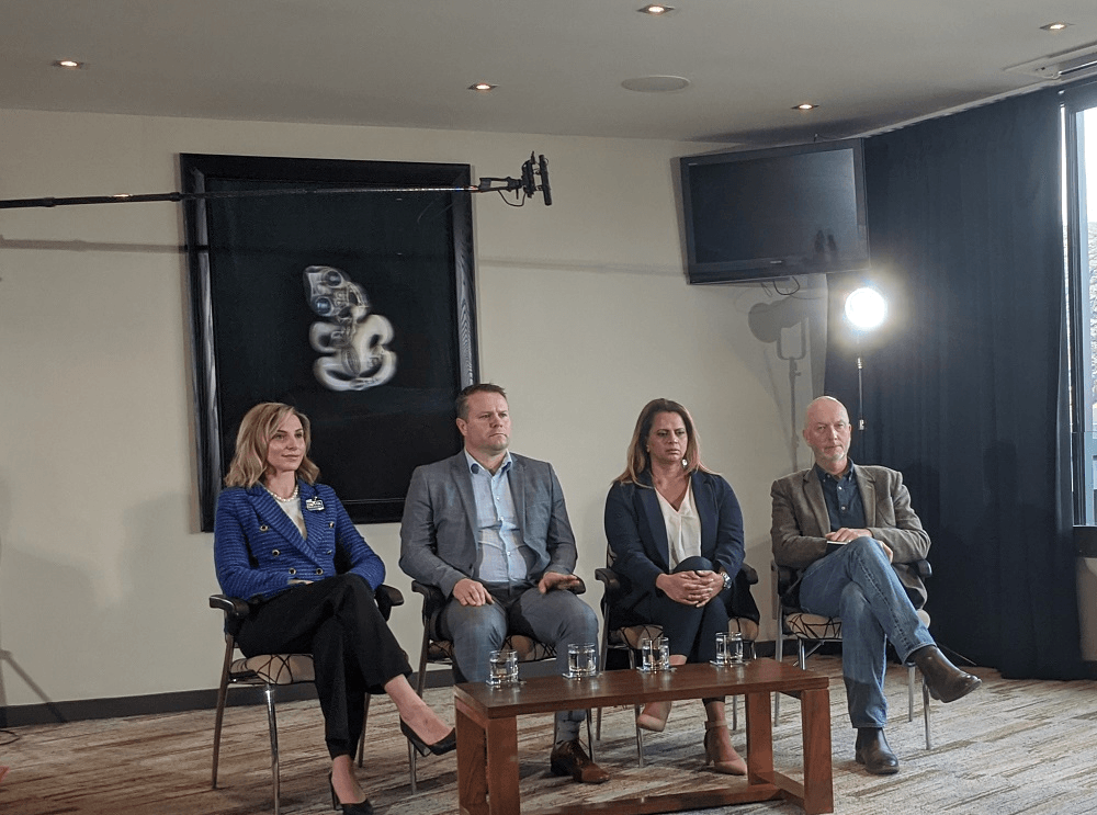 four people sitting in a row, white woman, white man in suit, brown woman in heels and blazer, bald white man in jeans.