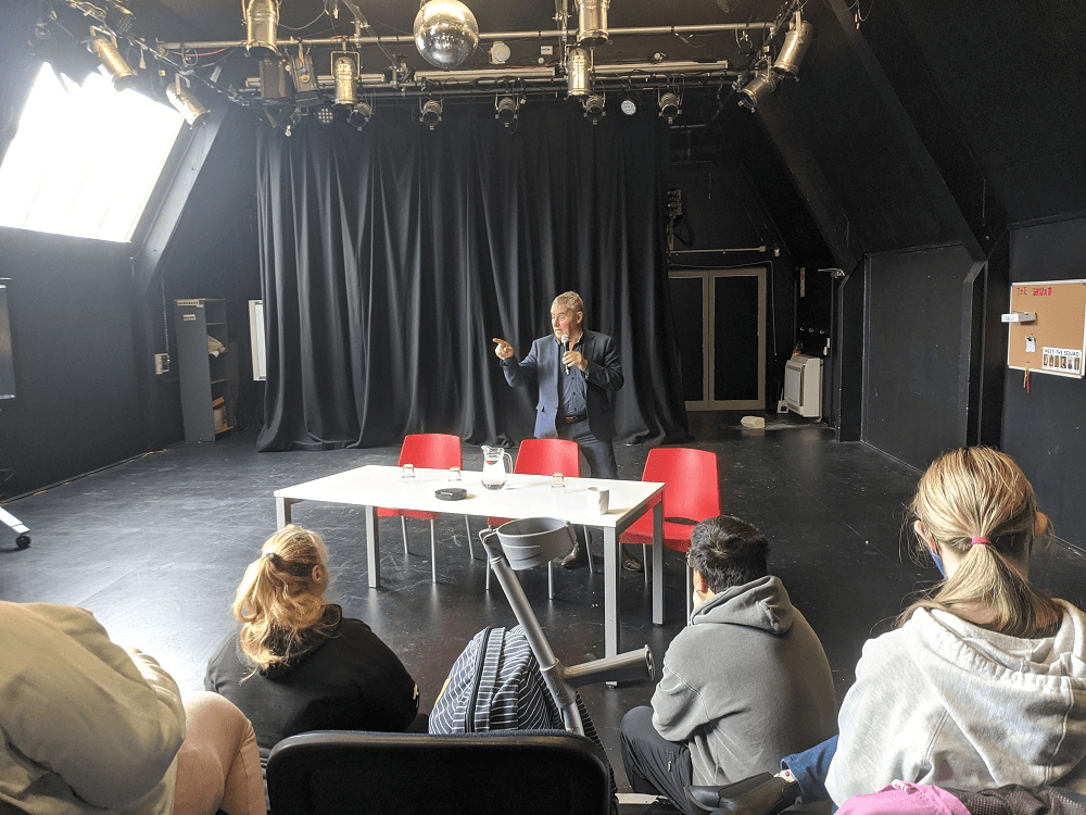 phil maguer in black room talking to people sitting on chairs who are looking at him