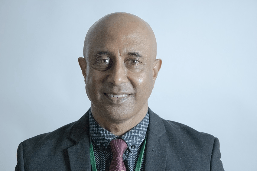 Lawrence naicker, a bald brown man wearing a suit, smiles in front of a blank wall