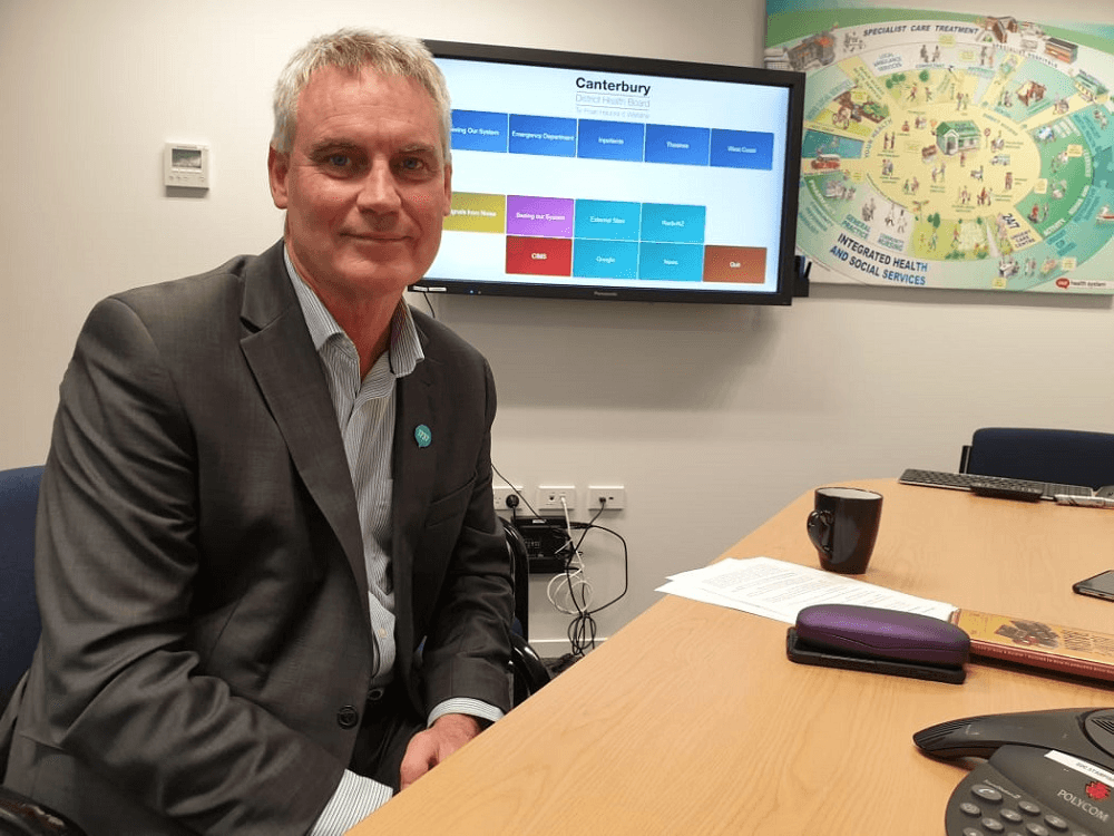 david meates in a suit in an office with a poster on the wall and paper on the table