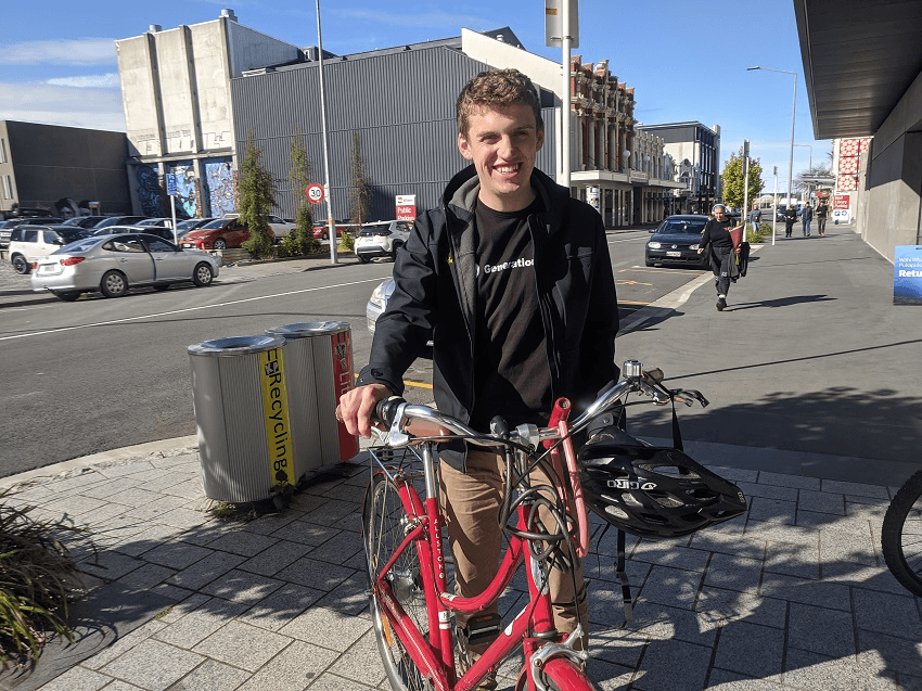 a sunny day in christchurch, nick a cheerful young white man wears a black tshirt that reads 'generation zero' and holds the handlebars of his bike