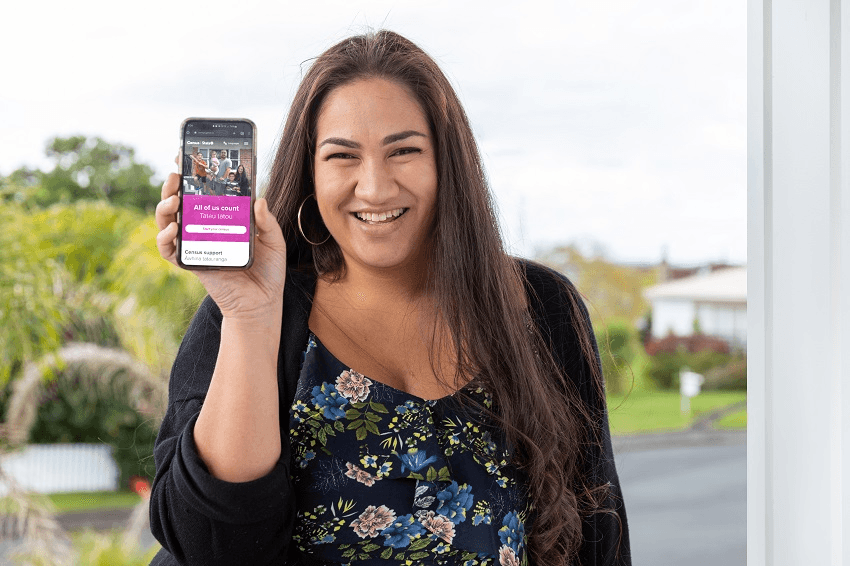 smiling woman with really great long brown hair that looks tidier than mine ever does has a big grin on her face and a phone displaying the trademark purple of the census website