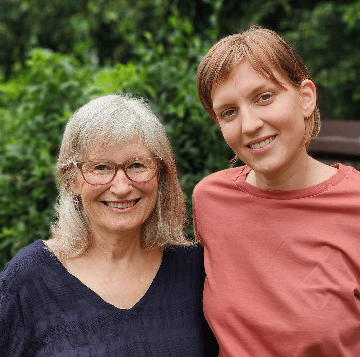 a grey haired white woman and her taller honey blonde daughter with dappled green leaves in the background