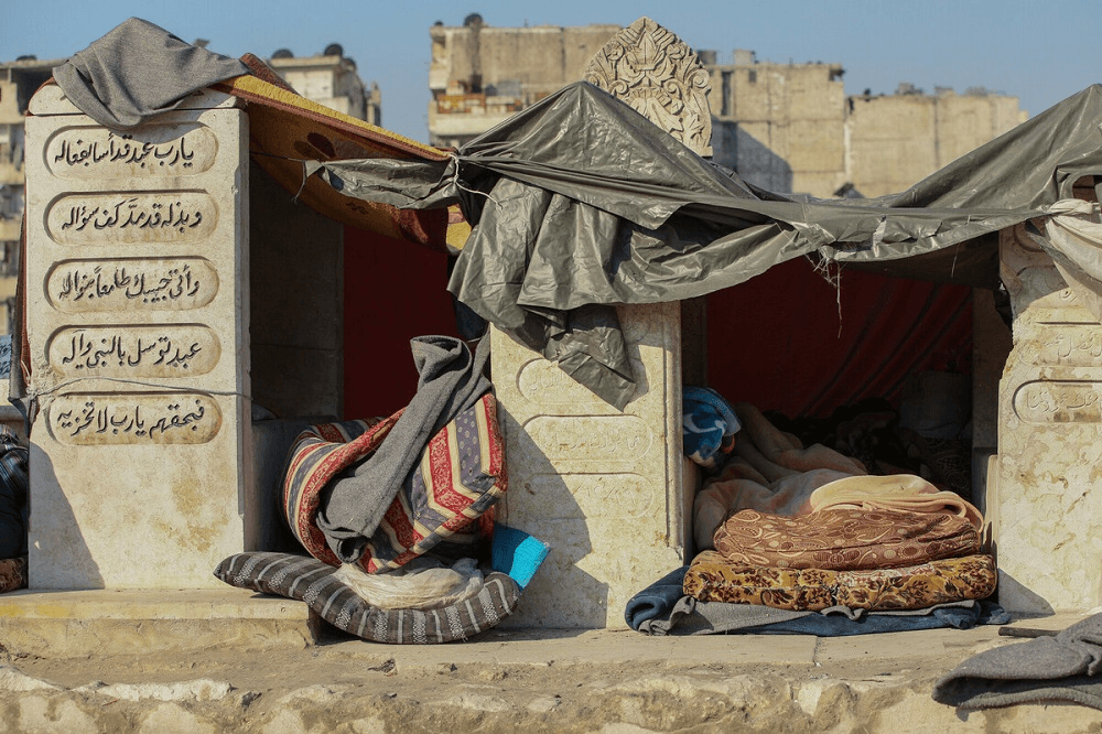 sunlight and the ends of two beds with a grey blue skky and bedding between two pillars, covered with plastic sheets and marked with Arabic writing 