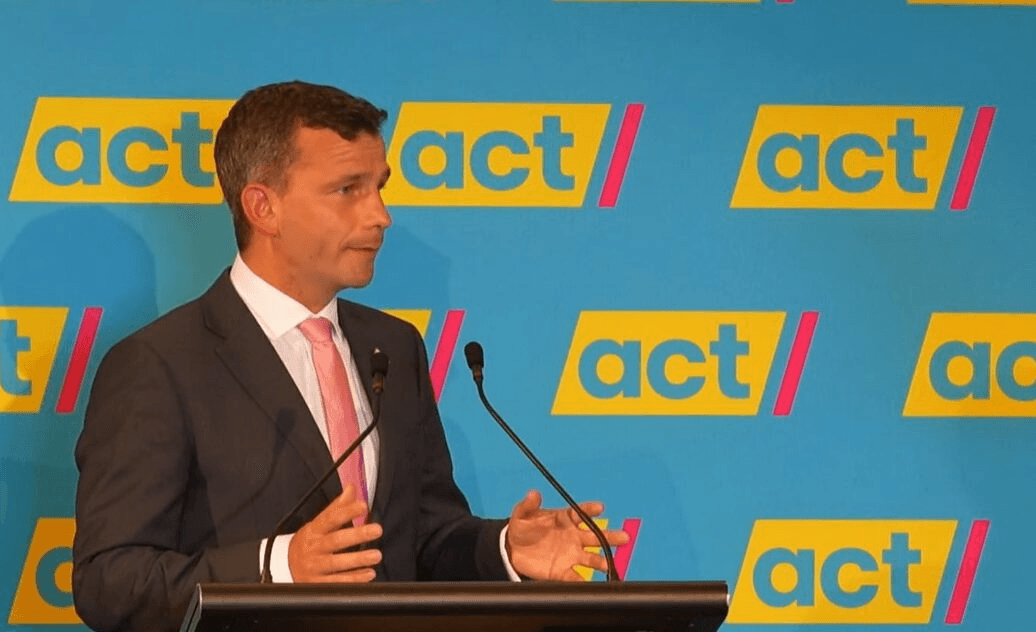 David seymour, a light-skinned man in a grey suit with a pink tie stands in front of a microphoone and the blue, yellow, and pink ACT logo