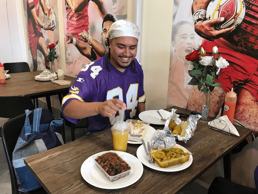 John-Paul Foliaki sits at a table with plates of Tongan food. He wears a bandana and a purple football jersey.