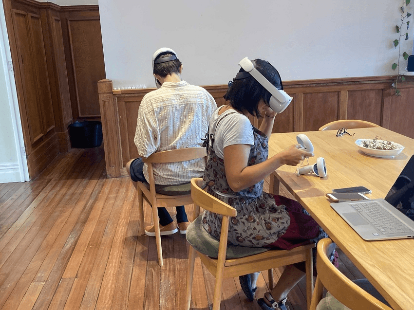 two people wearing white VR headsets face in opposie directions from each other, looking a bit confusing. It looks kind of glum and non social actually! one is a woman in a light grey tsuirt and block printed skirt and the other is a man in a white ish shirt.