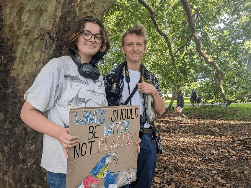 two young white people under a tree holding a sign that says 'waves should not be plastic'