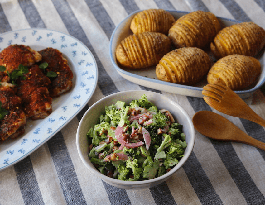 A bowl of raw broccoli salad next to a platter of six hasselback potatoes. The food is served on a striped tablecloth.