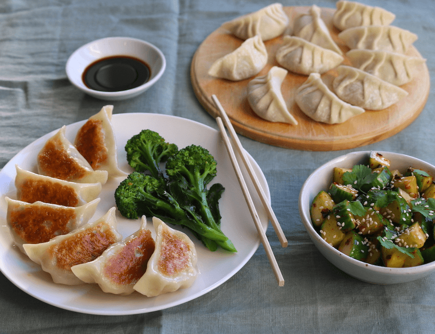A plate of fried dumplings beside a small pile of brocolli. Surrounding the plate are a small bowl of dipping sauce, a plate of freshly-made dumplings and a bowl of Chinese cucumber salad with coriander and sesame seeds.