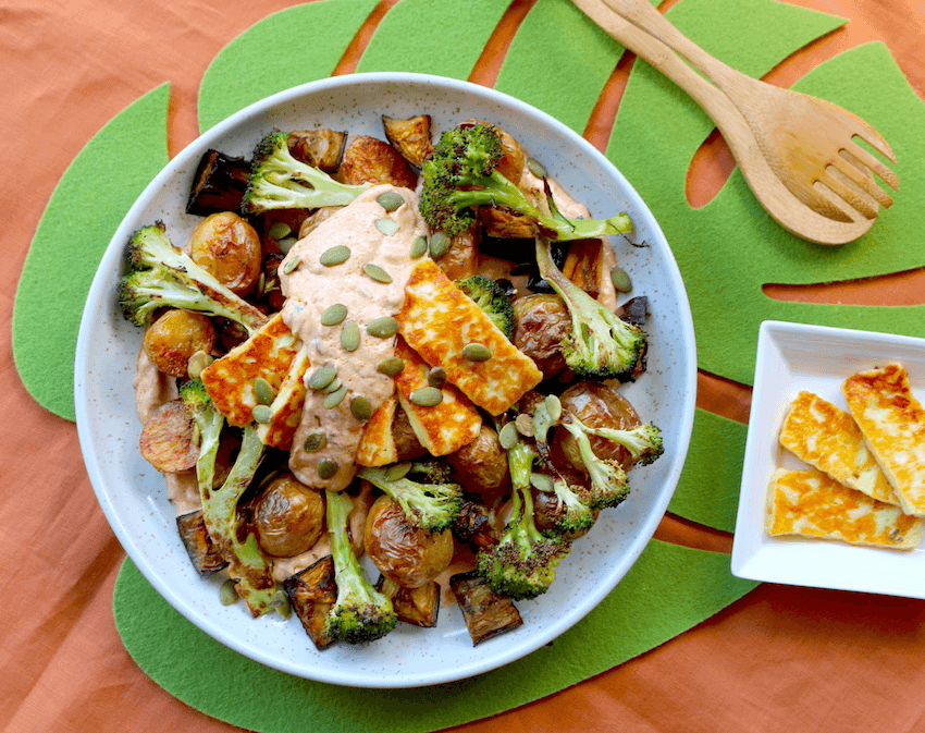 A bird's eye view of grilled haloumi atop a bowl of roasted potato, eggplant and broccoli. The plate is scattered with pumpkin seeds. The dish is displayed on top of a green monstera leaf placemat and a peach-coloured table cloth.