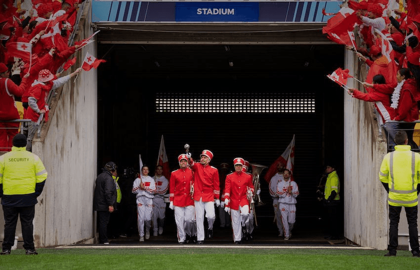 A still from the film Red, White and Brass. The band dressed in red marches onto the field surrounded by fans with Tongan flags.