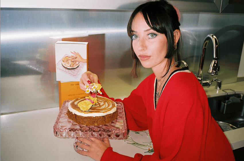The Caker Owner Jordan Rondel wears a red jumper and poses with an iced cake on a cake plate. She is standing in a kitchen with silver walls.