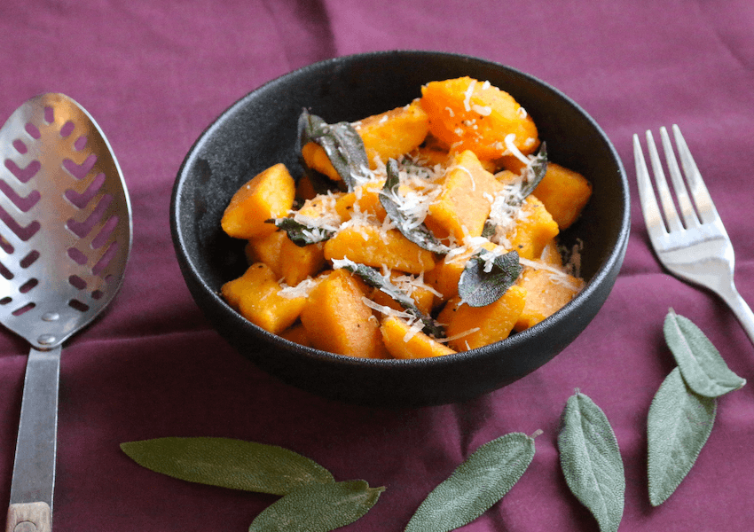A bowl of orange-coloured pumpkin gnocchi with grated parmesan and sage leaves on a purple table cloth.