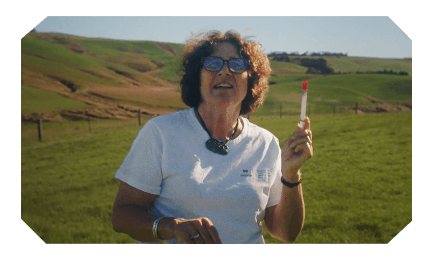 Dr Bev Lawton holds up a test tube in front of some rolling green hills of rural Otago